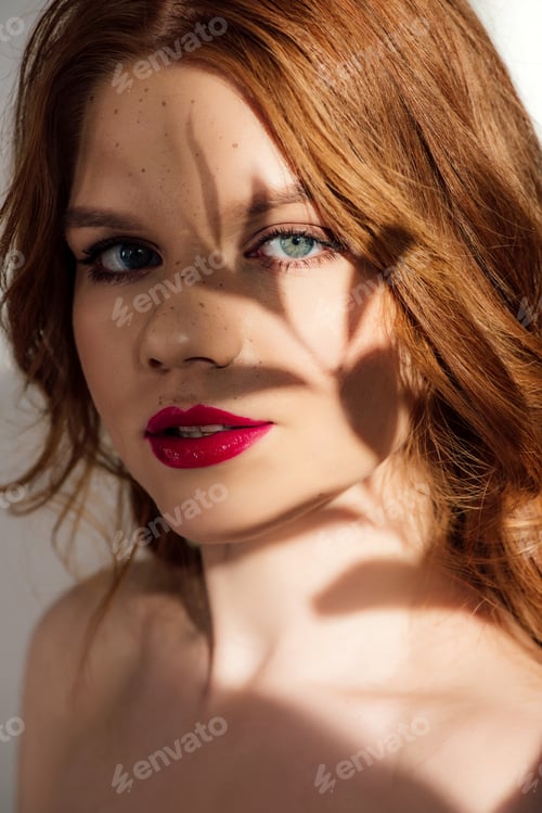 Preview: beautiful young redhead woman with red lips and shadow on face looking at camera