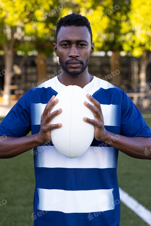 Preview: African American young male athlete holding a rugby ball on field outdoors, looking at camera