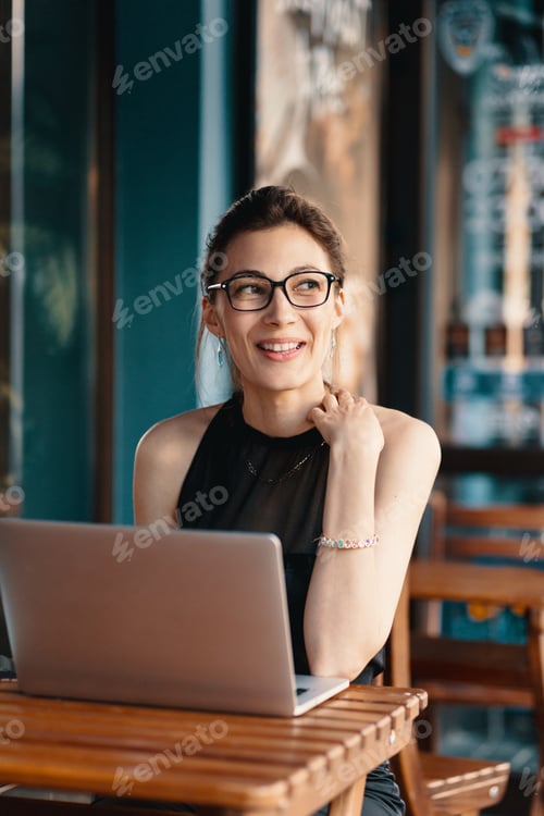 Preview: Refined business woman in glasses, sitting at table in cafe working at laptop