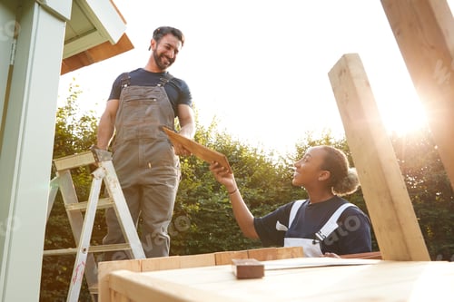 Preview: Male Carpenter With Female Apprentice Putting Roof On Outdoor Summerhouse In Garden