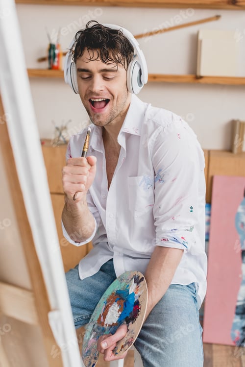 Preview: selective focus cheerful artist in headphones, singing with paintbrush while holding palette