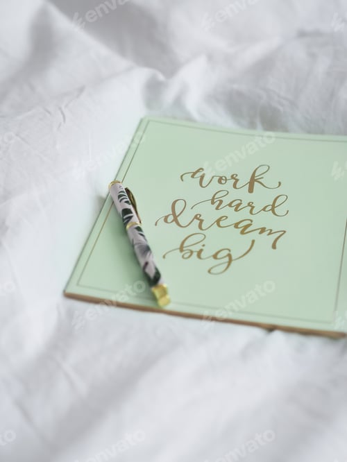 Preview: Vertical shot of a green notebook with an inspirational quote on a white cloth with a pen