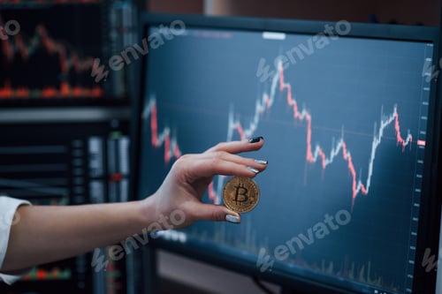 Preview: Front view. Woman's hand holding bitcoin in modern office with many monitors with graphs