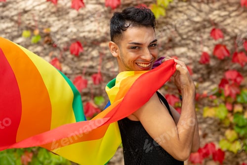 Preview: Portrait of happy biracial transgender man holding rainbow flag, with rocks and leaves in background