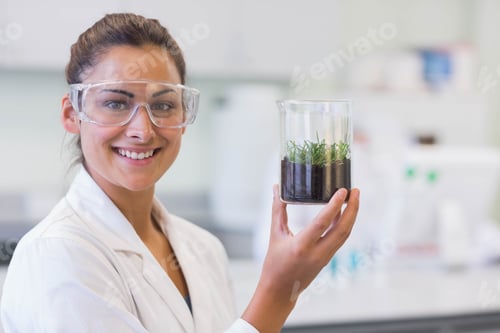 Preview: Female scientist wearing lab coat holding glass beaker with soil and seedlings at lab bench