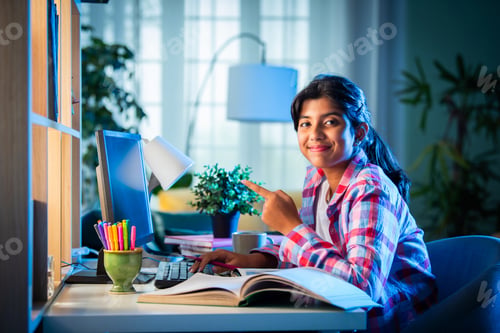 Preview: Asian Indian girl child studying at home on study table with computer, books, victory trophy