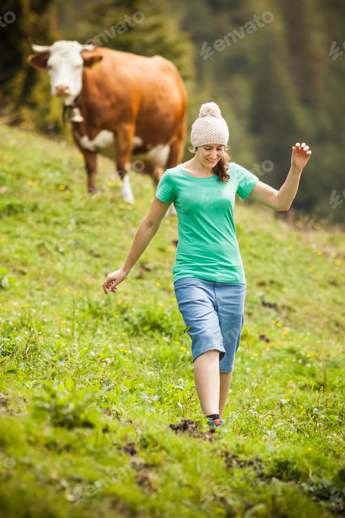 Preview: Young woman enjoying a leisurely walk in a lush green meadow with a cow observing in the background.