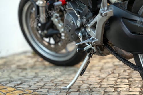 Preview: Detailed view of a motorcycle kickstand resting on cobblestone pavement in daytime