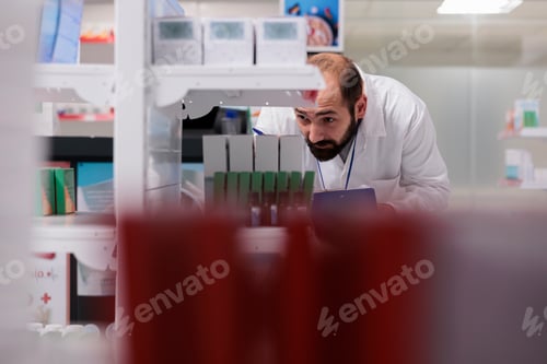 Preview: Pharmacist looking at shelves full with drugs packages doing inventory writing vitamins name on