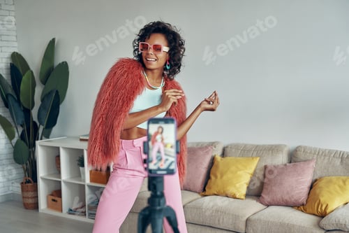 Preview: Attractive African woman in fluffy jacket creating her dancing video on phone camera at home