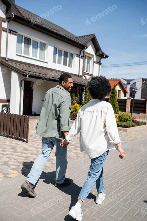 Preview: smiling african american couple holding hands while walking on sidewalk near houses on street