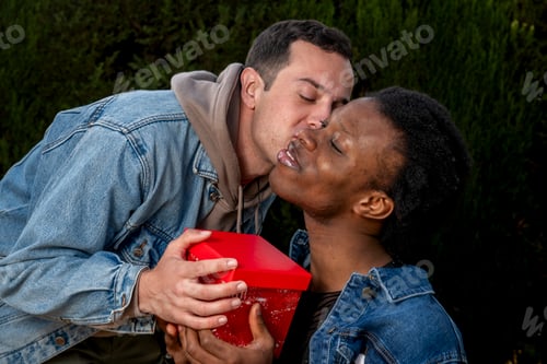 Preview: Man kissing woman holding red gift box outdoors