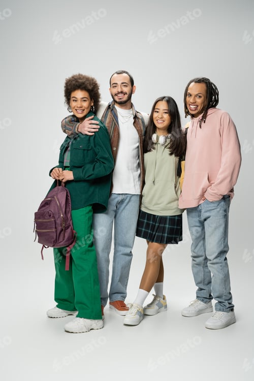 Preview: full length of stylish multiethnic students with backpacks looking at camera while standing on grey