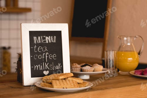 Preview: menu board, dishes with waffles and cakes, and jug with fresh juice on bar counter in coffee shop