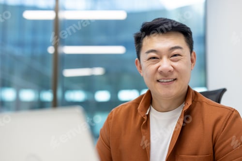 Preview: Asian businessman smiling in modern office setting with bright lights and large windows creating