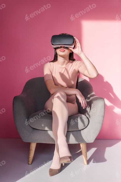 Preview: beautiful young woman using virtual reality headset and sitting in armchair on pink