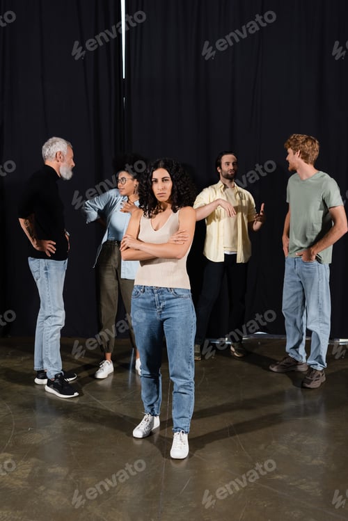 Preview: multiracial actress standing with crossed arms near interracial theater students and art director