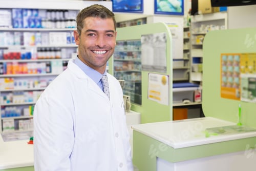 Preview: Smiling male pharmacist standing at counter in pharmacy, with medications and posters, copy space