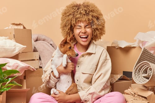 Preview: Positive curly haired woman poses with favorite pet exclaims loudly wears jacket surrounded by cardb