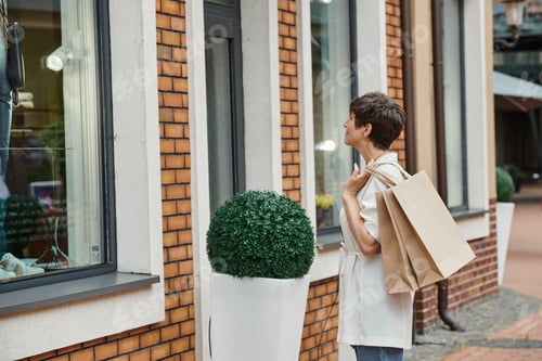 Preview: senior woman with short hair holding shopping bags and looking at window of outdoor mall, outlet