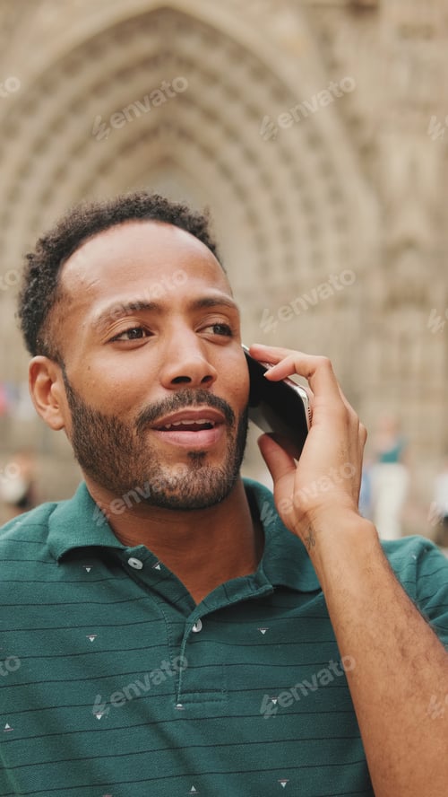 Preview: Happy young man sits on the steps of the Sagrada Familia in Barcelona, talking on smartphone