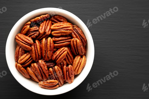Preview: Homemade Shelled Pecans in a bowl on a black surface, top view. Flat lay, overhead, from above.