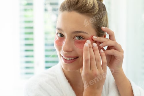 Preview: Happy caucasian woman in bathrobe putting gel mask under eyes in sunny bathroom