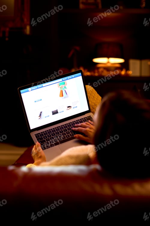 Preview: Rear View Of Woman At Home Sitting In Lounge At Night Shopping Online