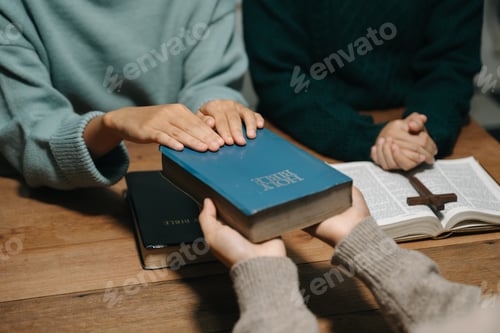 Preview: Group of Christians sit together and pray around a wooden table with blurred open Bible pages