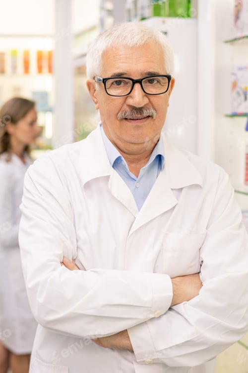 Preview: Aged drugstore worker standing against displays with medical goods and colleague