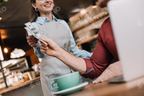 Preview: cropped view of freelancer with laptop giving cash to smiling waitress in coffee shop