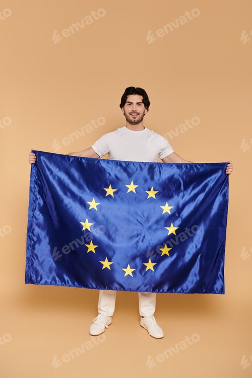 Preview: Young man confidently holding European flag against warm backdrop in a creative indoor setting