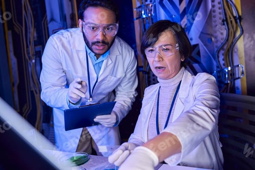 Preview: indian scientist with clipboard pointing at laboratory glass near colleague in innovative laboratory