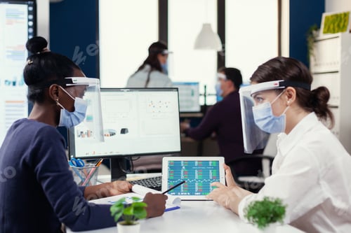 Preview: Multi ethnic employees with visor and protection mask sitting at desk