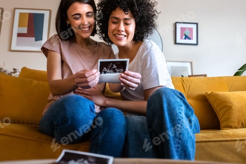Preview: Smiling multiracial lesbian pregnant couple looking baby ultrasounds at home sitting on the couch.