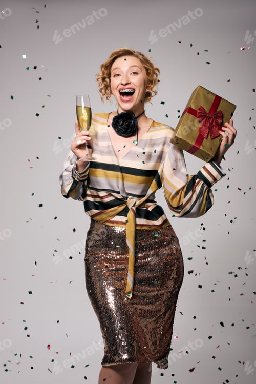 Preview: A cheerful young woman holds a champagne glass and a beautifully wrapped gift, exuding excitement
