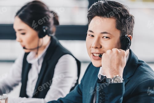 Preview: handsome young man in headset smiling at camera while working with female colleague in call center