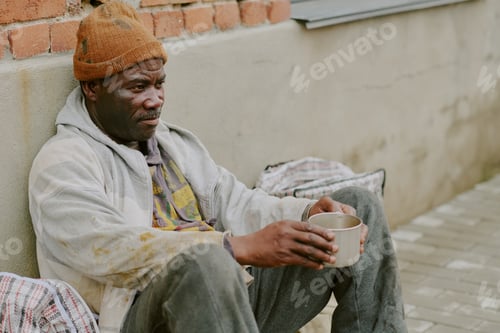 Preview: Homeless Black Man Sitting Against Wall Holding Cup Appearing Contemplative