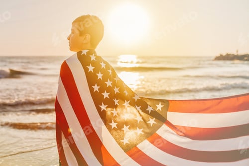 Preview: Back view candid authentic gen Z boy wrapped American flag looks at the sea on celebrate July 4th