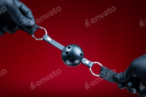 Preview: hands in black gloves holding a round gag in the mouth for erotic bdsm games