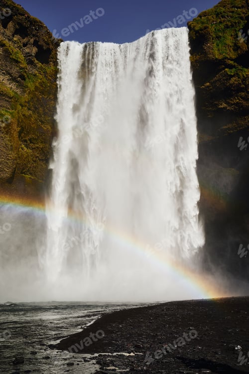 Preview: Stunning view of Skogafoss waterfall in Iceland with a vibrant rainbow arching across the mist.