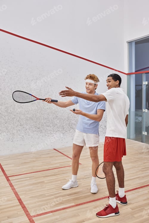 Preview: happy interracial male friends pointing with racquets and looking away inside of squash court