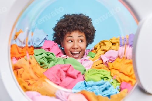 Preview: Head of funny woman with cheerful expression stuck in heap of mutlicolored laundry blue background.