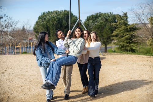 Preview: Happy young adults having fun pushing friend on swing in playground