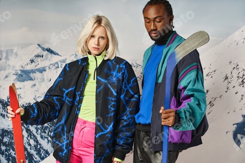 Preview: attractive interracial couple in vibrant warm outfit holding skis and hockey stick, winter concept