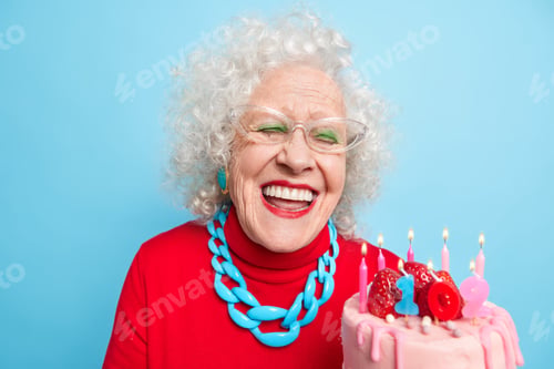 Preview: Positive aged woman with grey curly hair smiles broadly has white perfect teeth holds cake celebrate