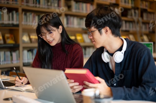 Preview: Glasses asian man student looking at woman friend writing in notebook while sits at table in library