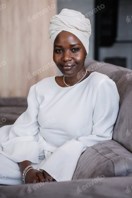Preview: Young African woman in white dress and turban sitting in couch looks at camera confidently