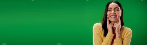 Preview: young brunette woman with closed eyes touching cheeks and smiling isolated on green, banner