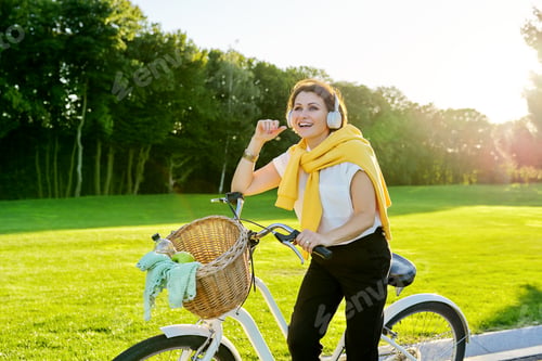 Preview: Beautiful happy middle-aged woman in headphones with bicycle in park